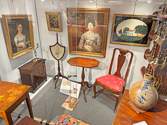 Behind glass doors is an area that Hillier arranges into vignettes. He calls it the “museum case.” The oil on canvas portrait of a young girl with a basket of flowers (left) is tagged $3250; the pole screen with embroidery is tagged $1050; the portrait of a man with a tricorn hat (above) is tagged $595; the Queen Anne candlestand (center) with an octagonal top, North Shore, Massachusetts, is tagged $800; the American school portrait by an unknown artist of a woman seated at a worktable creating hair jewelry is tagged $2450; the walnut side chair, Massachusetts, 1740-1760, is tagged $4985; the schoolgirl watercolor painting of George Washington’s Mount Vernon is tagged $2450; and the Maine grain-painted hanging shelf, circa 1822, is tagged $6250. “I had this built years ago because I wanted changing exhibits,” said Hillier. It changes every month.