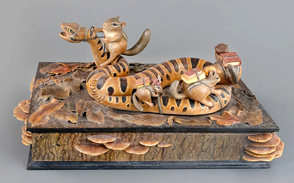 Frank S. Finney (b. 1947) carved the group of three small chipmunks riding atop a rattlesnake while a larger chipmunk holds the reins, appearing to drive the snake, all of which are set out on a book (or a box) with a single drawer, 25