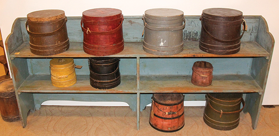 Bucket bench in blue paint, $950; firkins on the upper shelf, from left: brown, $265; red, $275; gray, $295; and brown, $225; firkins on the lower shelf, from left: mustard, $295; black, $255; and red, $130; on the floor: kerosene container, $175; and green firkin, $285 from Eric D. Nichols of Tavern Creek Antiques, Portland, Missouri.