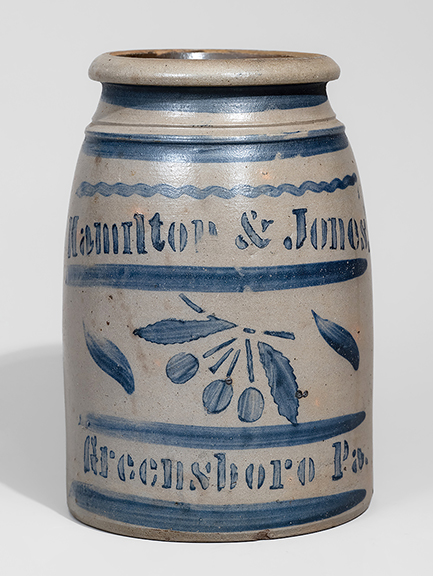 This western Pennsylvania, circa 1870, stoneware canning jar, 9 3/8” tall, with cobalt freehand and stenciled cherry decoration, stenciled “Hamilton & Jones. / Greensboro Pa.,” sold for $7800 (est. $2500/3500).