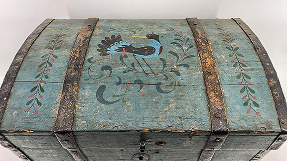 The lid of this diminutive blue-painted dome-top wooden chest is decorated with a large stylized bird, flanked by floral sprigs. The chest is trimmed with wrought-iron straps. The two medial straps are not only decorative; they are the tails of the chest’s hinges and completely surround the chest. There is an internal lidded till. The forged handles are attached with snipe-style fasteners. The 10½