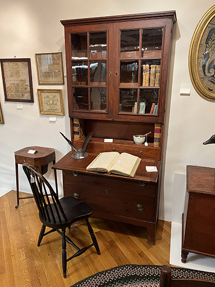 The mid-19th-century two-piece red secretary from Woodstock, Vermont, has original old glass (some damaged) and was $3250 from Pewter & Wood Antiques, Enfield, New Hampshire, and Cave Creek, Arizona. On it were an 1840s leather-bound ledger, Salisbury, New Hampshire, or Massachusetts, $95; a 19th-century mortar and pestle, $95; and a New England 19th-century pewter inkwell with a porcelain insert, $289. The Windsor chair in black paint was $375.