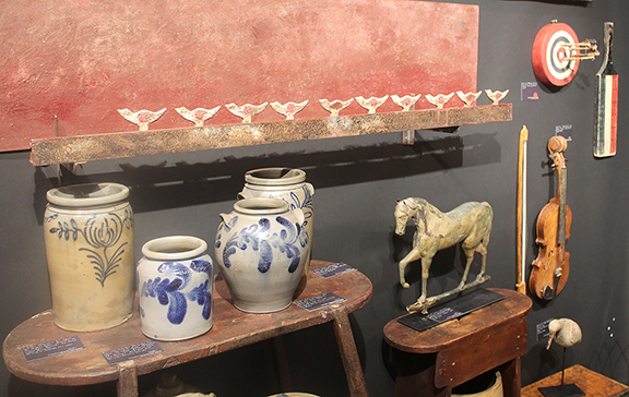 The booth of Christopher and Bernadette Evans Antiques, Waynesboro, Virginia, was filled with a variety of traditional folk art and Americana. The early 20th-century shooting gallery target and backstop was priced at $1750. The four decorated stoneware crocks are all by 19th-century Alexandria, Virginia, makers and were priced (from left to right) at $495, $1750, $1650, and $705. The zinc- and copper-body horse-form weathervane in as-found condition was marked $1750. The cribbage board was $550, and the 20th-century matchstick violin and bow were $895.