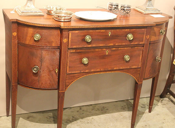 This diminutive six-leg sideboard has a confirmed attribution to Concord, Massachusetts, cabinetmaker, William Monroe/Munroe (1778-1861). Entries in his daybook date this piece to 1806-08. The sideboard features extensive inlay, drawer banding, and lightwood escutcheons. The forward edges of the top feature a “running arrow” inlay. There are two distinctive features of the case construction: bold “sash corners” and double-tapered legs below the cuff area. The sideboard was offered by Paul A. Thien of Firehouse Antiques Center, Galena Maryland, for $18,000.